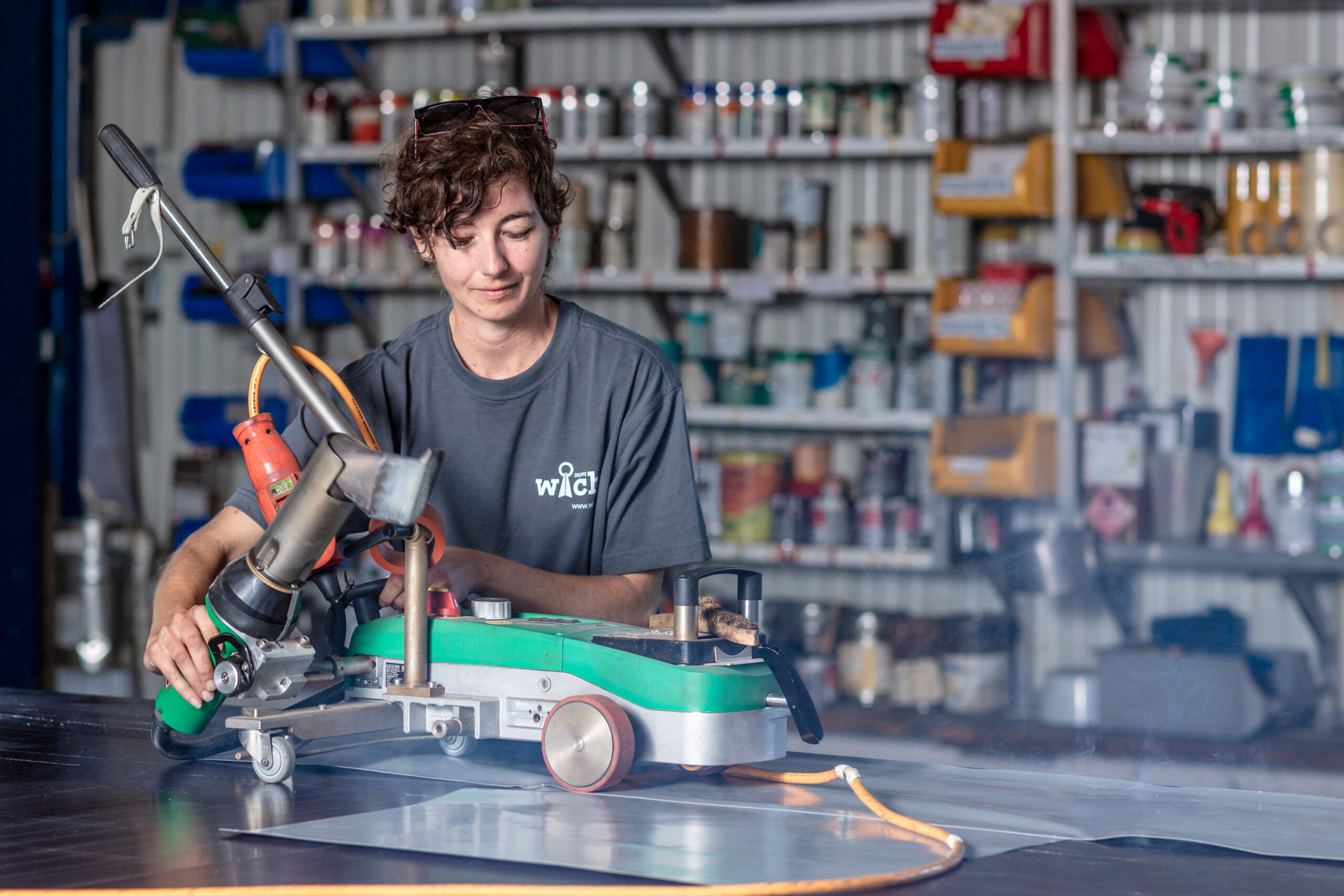 Eine Person bedient ein großes grünes Elektrowerkzeug auf einer flachen Metallfläche in einer Werkstatt. Im Hintergrund befinden sich Regale mit verschiedenen Werkzeugen und Behältern. Die Person trägt ein dunkles T-Shirt und hat einen konzentrierten Gesichtsausdruck.