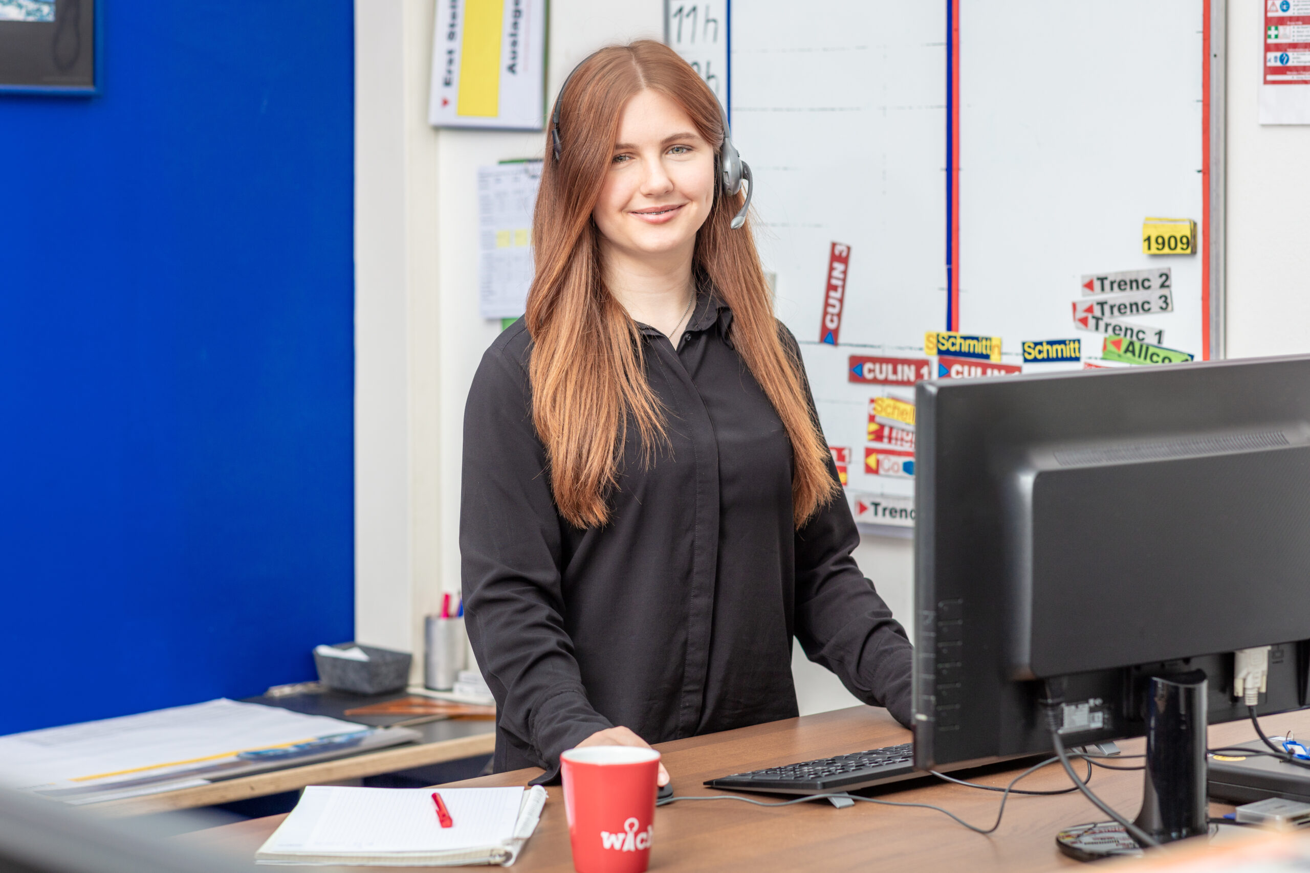 Eine Person mit langen Haaren, einem schwarzen Hemd und einem Headset steht an einem Schreibtisch mit einem Computer. Auf dem Schreibtisch stehen eine rote Tasse und ein Notizbuch. Im Hintergrund sind ein Whiteboard und eine blaue Wand zu sehen.