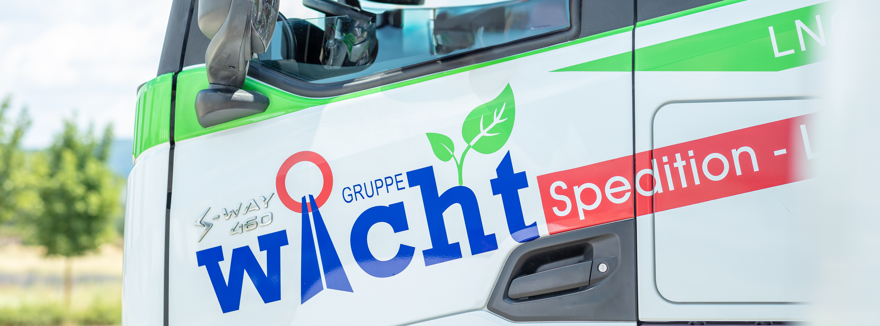 Nahaufnahme einer LKW-Seite mit den großen Buchstaben „Wicht Gruppe“ und „Spedition-Logistik“. Das Design enthält Blättergrafiken, die auf ein umweltfreundliches Thema hindeuten. Der LKW ist grün und weiß, mit verschwommenen Bäumen im Hintergrund.